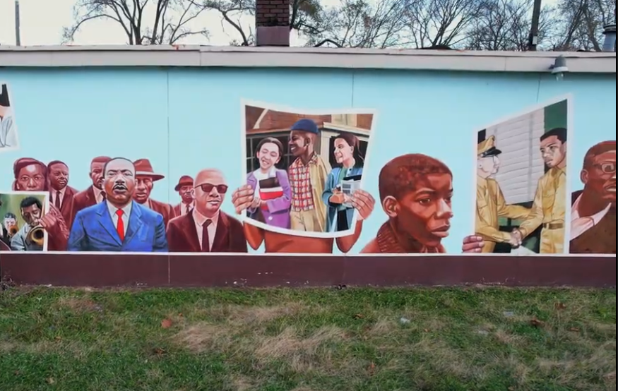 A mural on an outdoor wall depicting notable civil rights figures, community members holding up photos, and scenes symbolizing unity and struggle. The background shows a grass area and leafless trees.